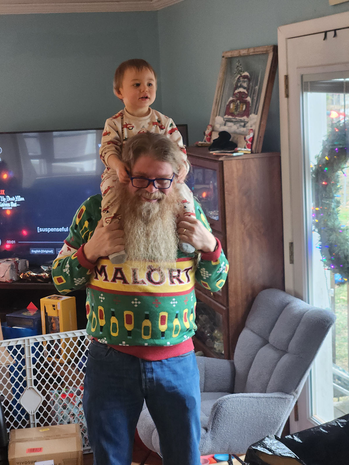 Photo of Jim, Damian's dad, a stocky man with glasses, a low pony, and a wide and long beard, smiling with mirth. On his shoulders is Cayden, a gleefully babbling toddler who is tryig to grab his glasses.