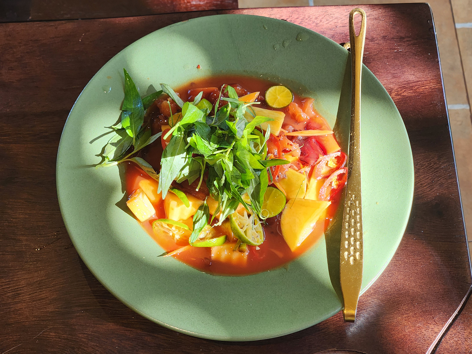 An image of a salad in a green ceramic bowl on a wooden table dappled with sunlight. The salad consists of orange mango chunks, small green rounds of kumquat, red slivers of chili, and red chunks of tree gum, sitting in a red dressing and topped with a pile of greenery.