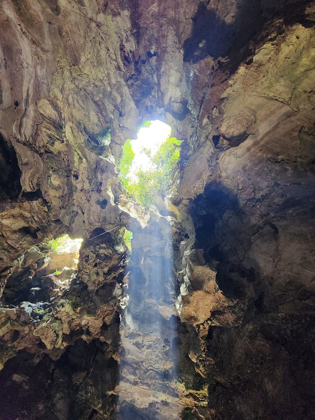A photo taken from inside a cave, aimed up at a hole to the surface through which a dramatic sunbeam shines through, illuminating the cave.