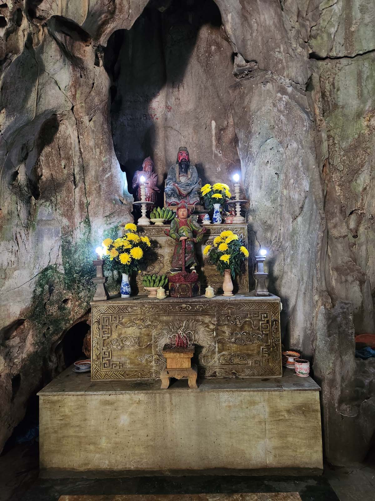A multi-tier stone altar with three small human statues on it. The top statue is of a man with green robes and a red face. On the altar there are vases with yellow flowers and plates of small green bananas.