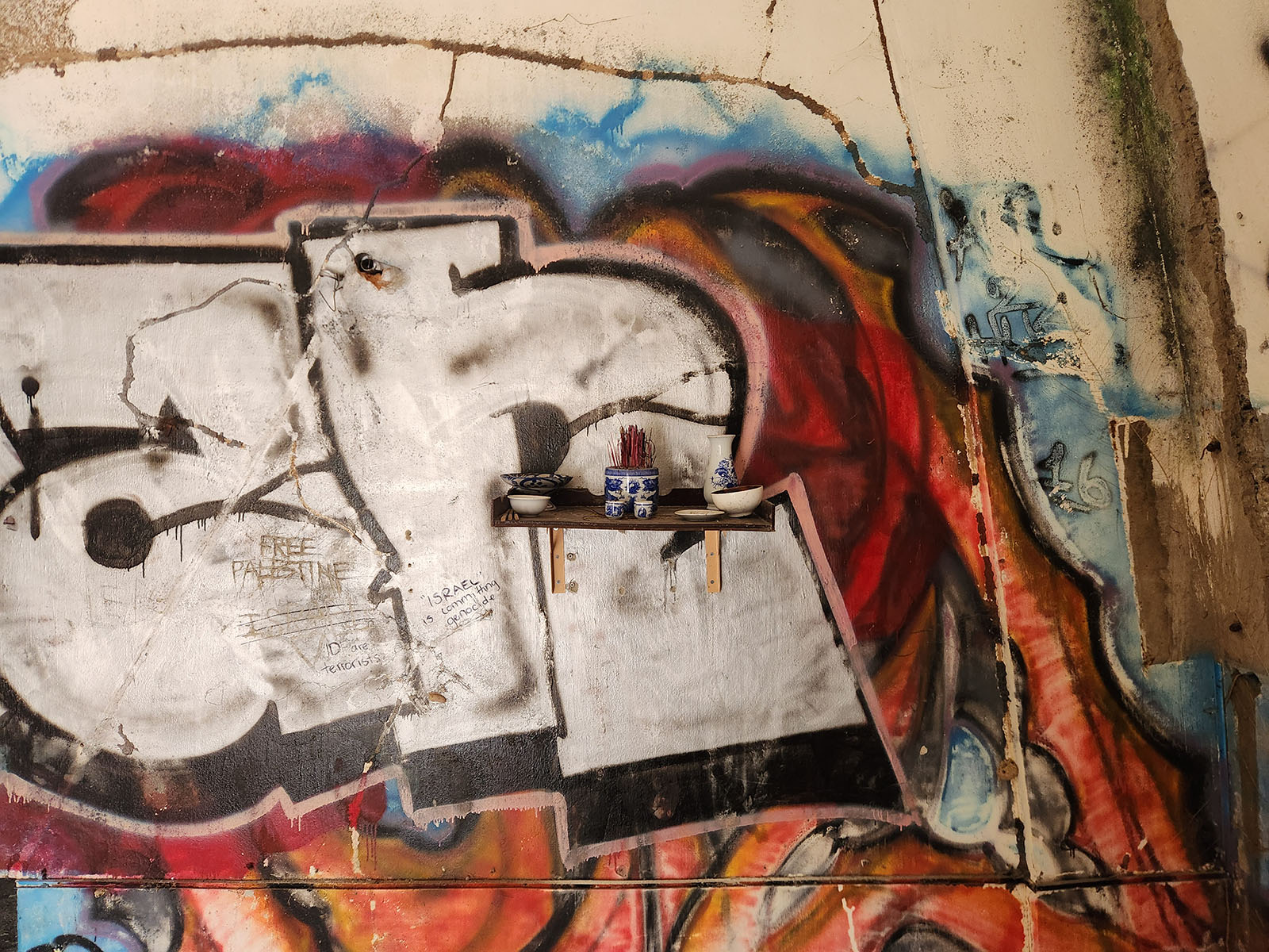 A wall covered in graffiti. A small shelf is mounted to the wall, atop which are placed small bowls and a pot of incense.